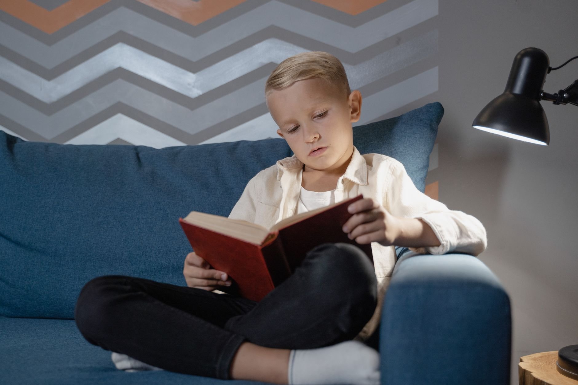 A young boy reading a book on a comfortable couch under warm lamp light